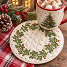 A round quilted Christmas-themed coaster in cream with green wreath embroidery and red berry accents, shown on a rustic wooden surface with a matching red-and-white plaid cloth, a decorated mug, pinecones, and holiday greenery.