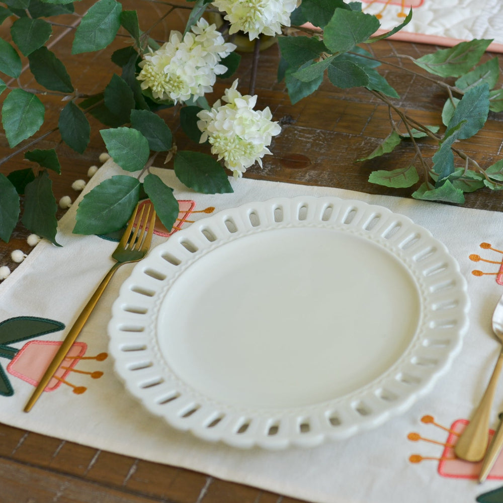 White plate on a decorative placemat with cutlery and flowers on a wooden table