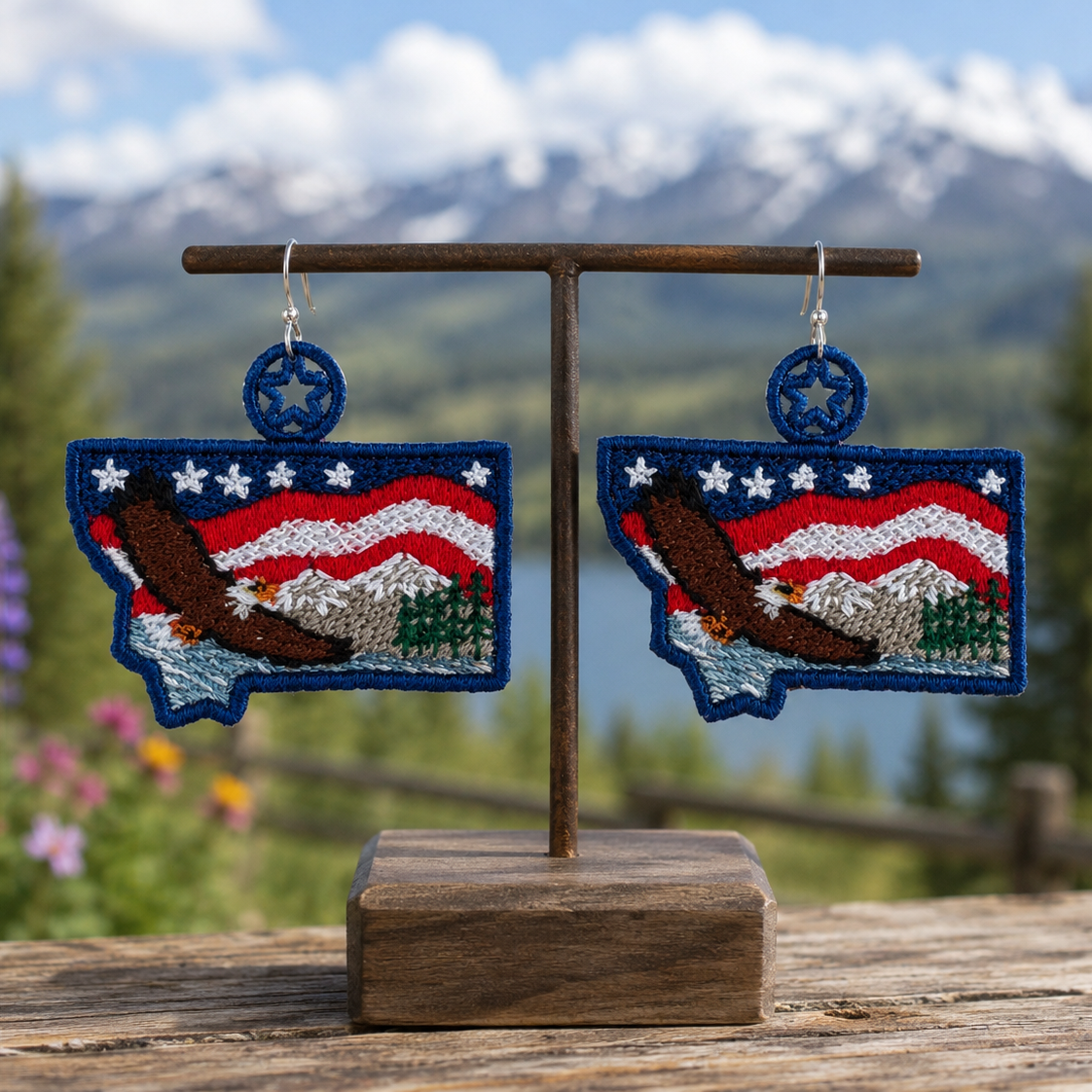 Pair of earrings shaped like the state of Montana with American flag design on a wooden stand against a mountainous background.