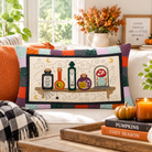 A square decorative throw pillow with a Halloween-themed patchwork border and central illustration of potion bottles, a spiderweb, and a mushroom, styled on a white sofa in a cozy autumn living room setting.