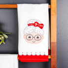 A white kitchen towel featuring an embroidered cartoon granny face with glasses, white hair, and a red bow, hanging on a wooden ladder against a dark gray wall.