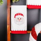 A white Christmas kitchen towel with an embroidered Santa face and red hat, hanging on a wooden ladder next to a red and white striped ornament against a dark gray wall.