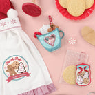 Collection of Christmas-themed kitchen items including an apron, cookie cutters, and decorative items on a pink background.