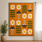 A large quilted wall hanging or tapestry featuring a mid-century modern floral and mushroom pattern on an orange background, styled in a room setting with a potted plant and white curtains.