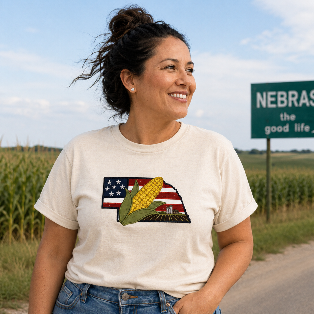 Woman wearing a t-shirt with a corn and American flag design, standing in front of a Nebraska sign.