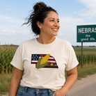 Woman wearing a t-shirt with a corn and American flag design, standing in front of a Nebraska sign.
