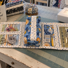 A fabric sewing organizer case laid open on a green cutting mat, holding scissors, thread, pins, ruler, and small accessories. A sewing machine, fabric stack, and sewing supplies are visible around it in a real craft workspace, so this is Lifestyle rather than Product/Packaging.