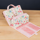 A floral travel toiletry bag with a matching zippered pouch, displayed open on a light wood tabletop. The toiletry bag features a pink handle, a pink adjustable strap with a buckle, and a white fabric body decorated with a pink and blue floral pattern.