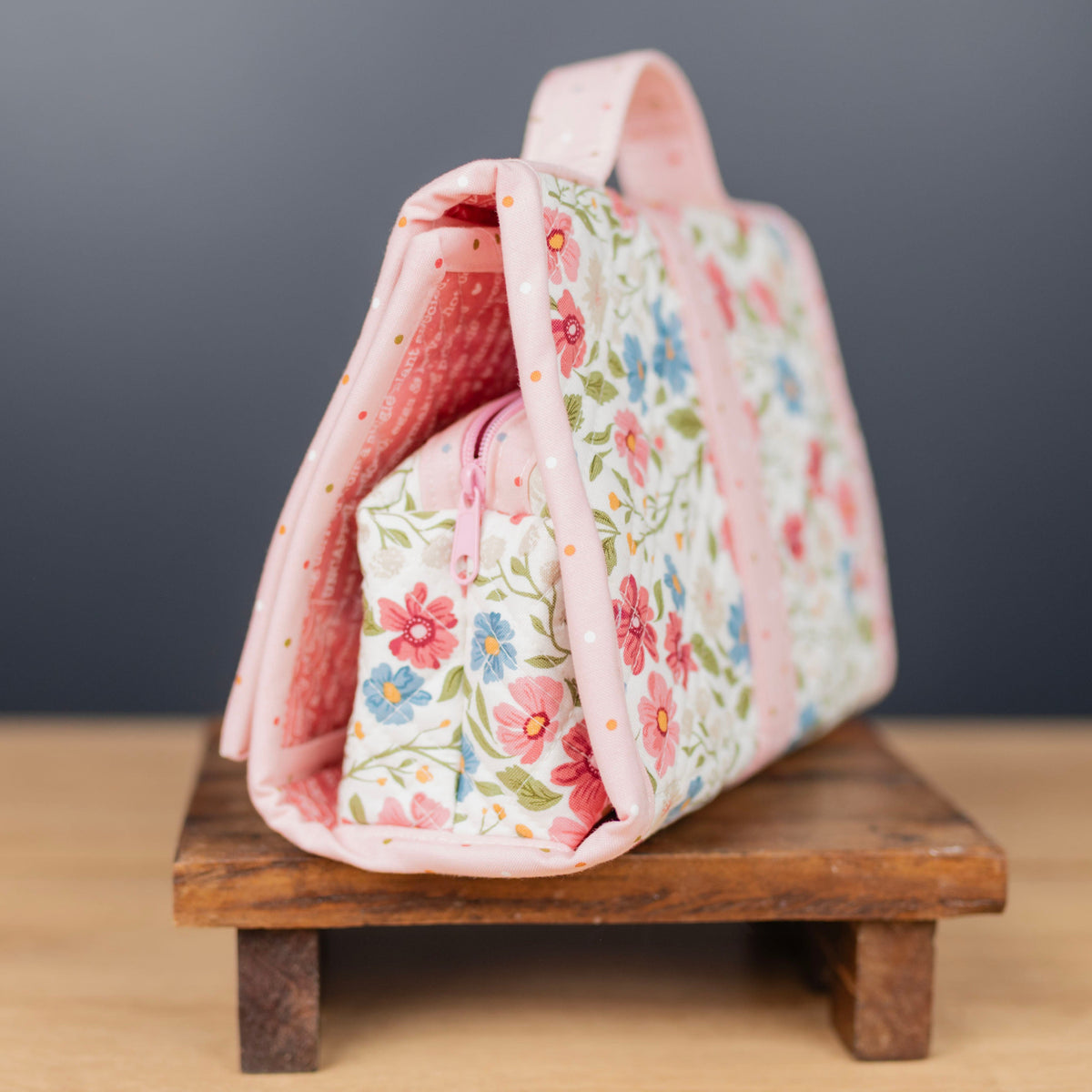 A pink floral fabric organizer bag with a matching zippered pouch, displayed on a small wooden stand against a dark gray background.