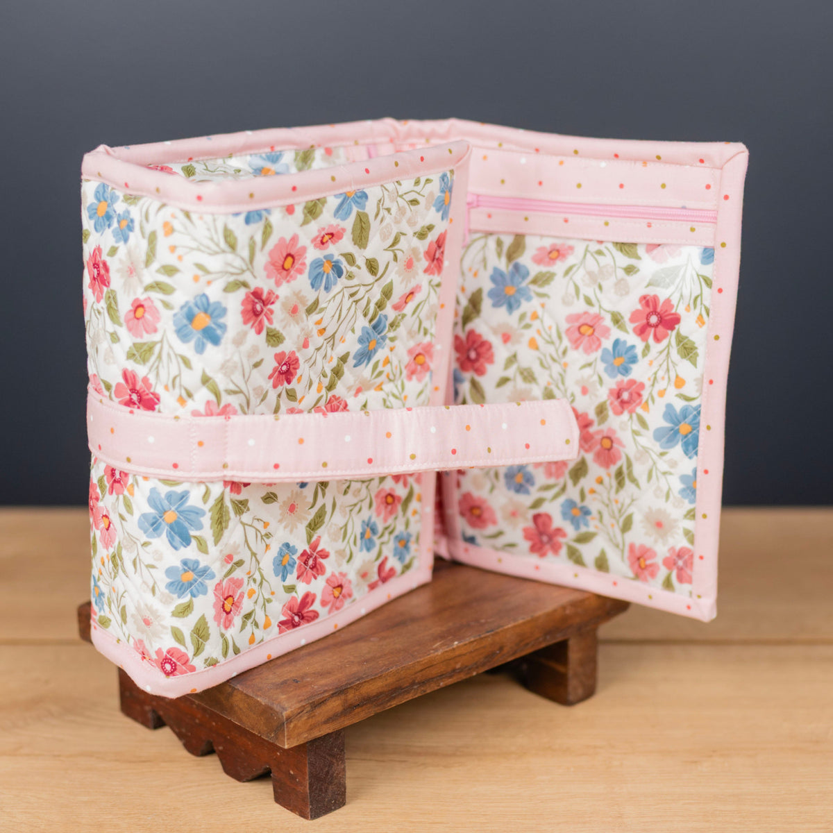 A fabric organizer pouch with a pink polka-dot trim and a floral print featuring pink, blue, and green flowers on a cream background, displayed open on a small wooden stand against a dark gray background.