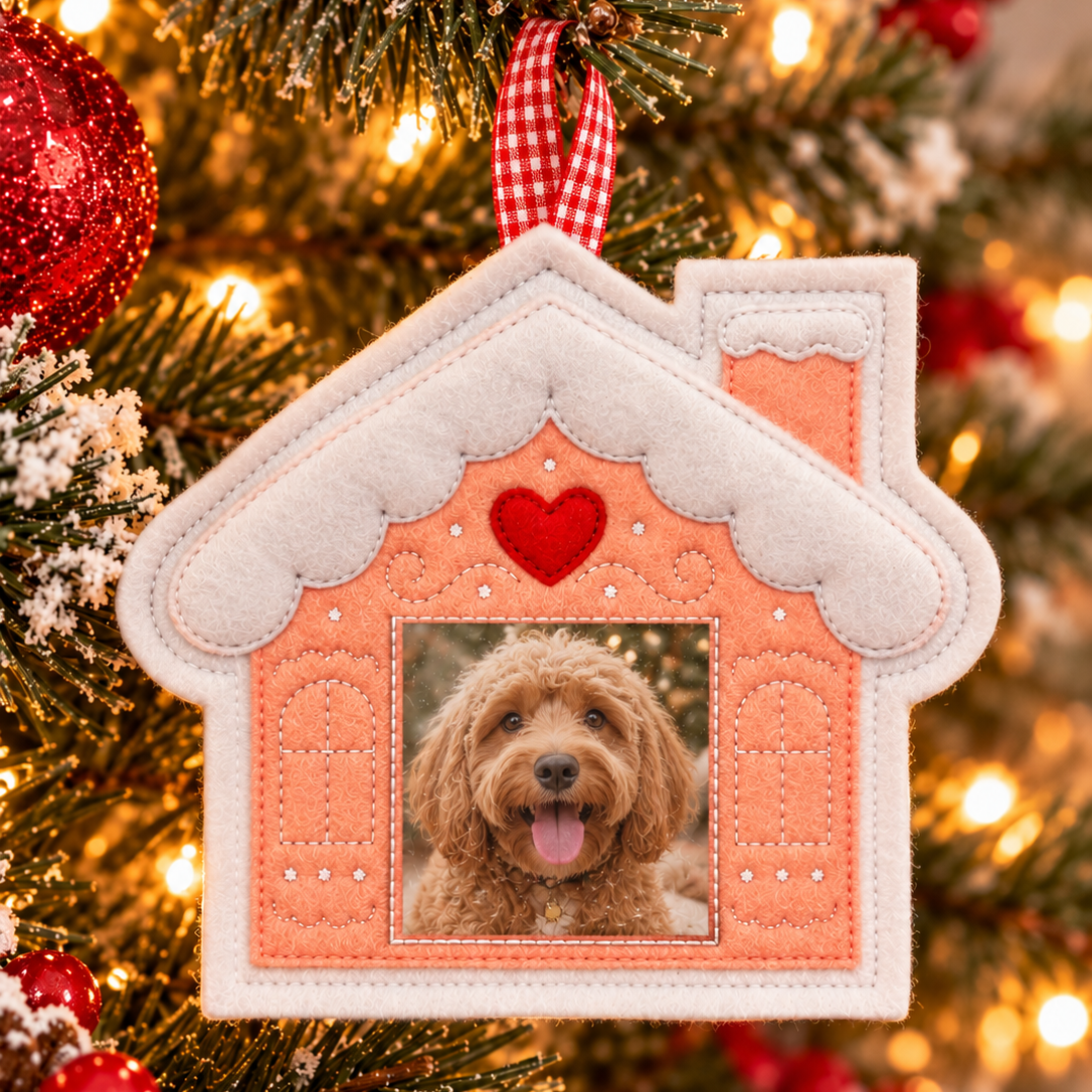A house-shaped Christmas ornament hanging on a decorated Christmas tree. The ornament has a peach-pink center, white faux-snow trim, a red heart near the top, and a square photo window showing a fluffy dog.