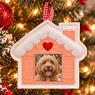 A house-shaped Christmas ornament hanging on a decorated Christmas tree. The ornament has a peach-pink center, white faux-snow trim, a red heart near the top, and a square photo window showing a fluffy dog.