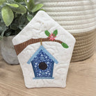 Decorative quilted birdhouse with embroidery on a wooden surface with plants in the background