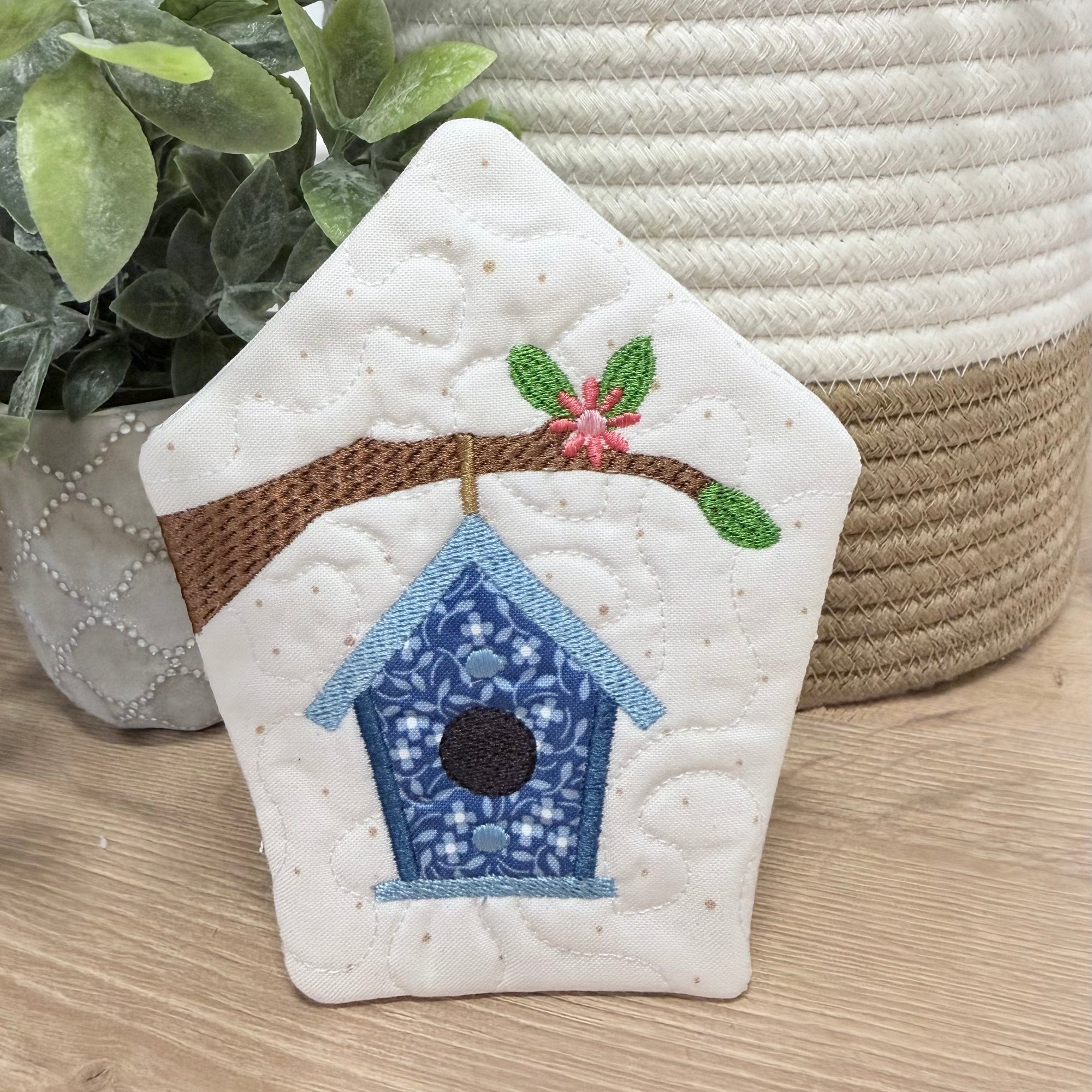 Decorative quilted birdhouse with embroidery on a wooden surface with plants in the background