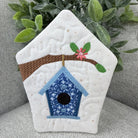 Quilted patch with a birdhouse design on a gray surface with greenery.