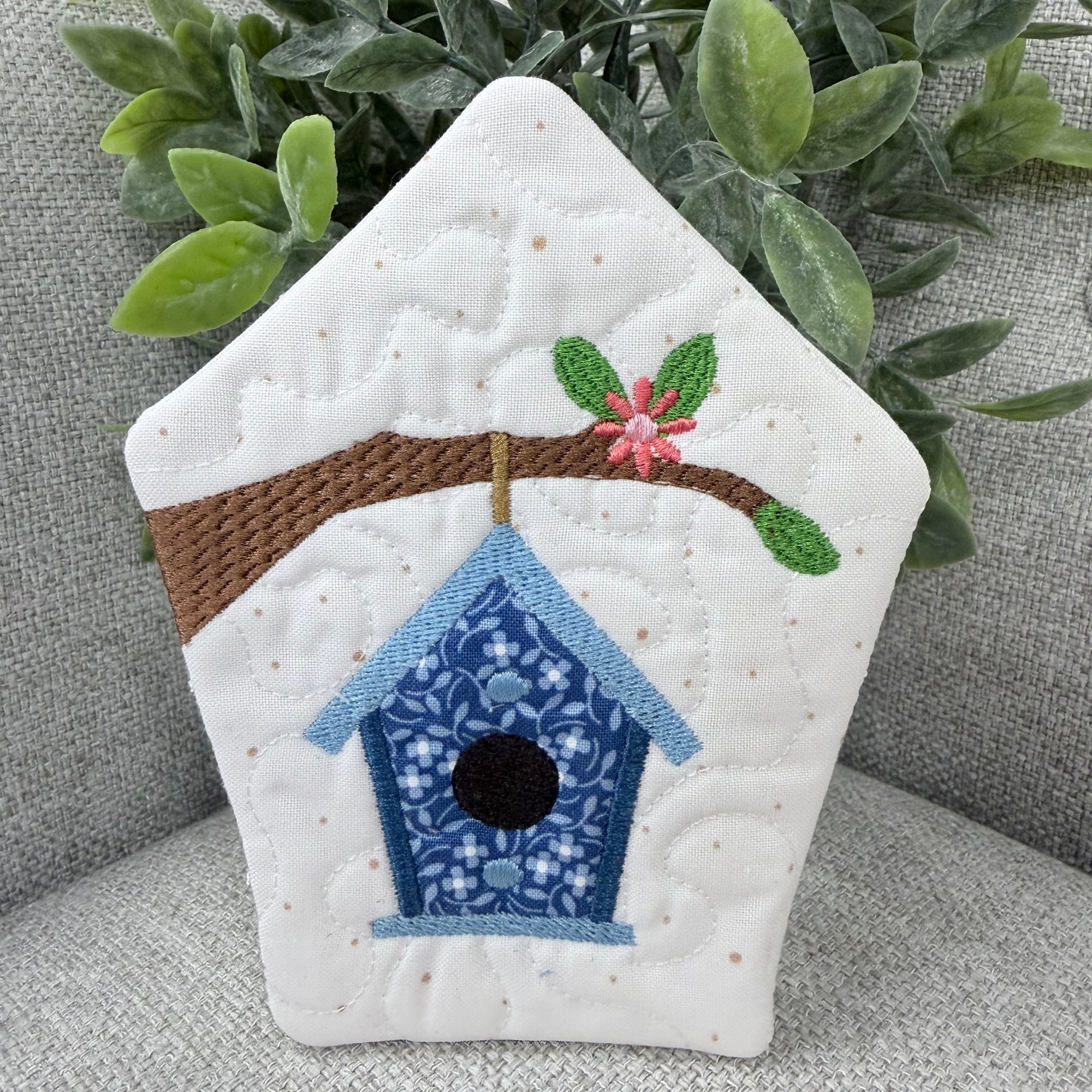 Quilted patch with a birdhouse design on a gray surface with greenery.