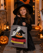 A young girl in a black witch costume and hat holds a gray fabric tote bag decorated with embroidered ghosts, a witch hat, books, and autumn leaves, standing in front of a warmly lit Halloween house porch with pumpkins.