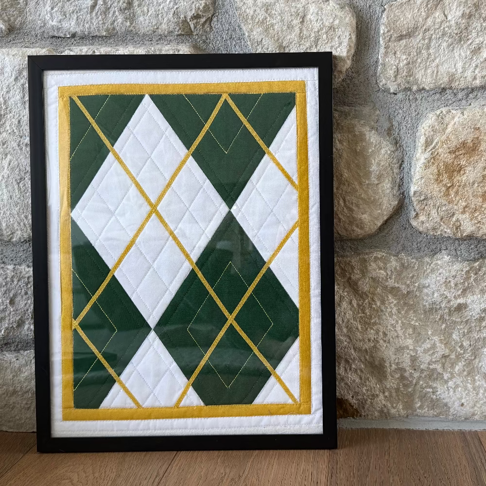 Framed quilt with green, white, and gold pattern against a stone wall.