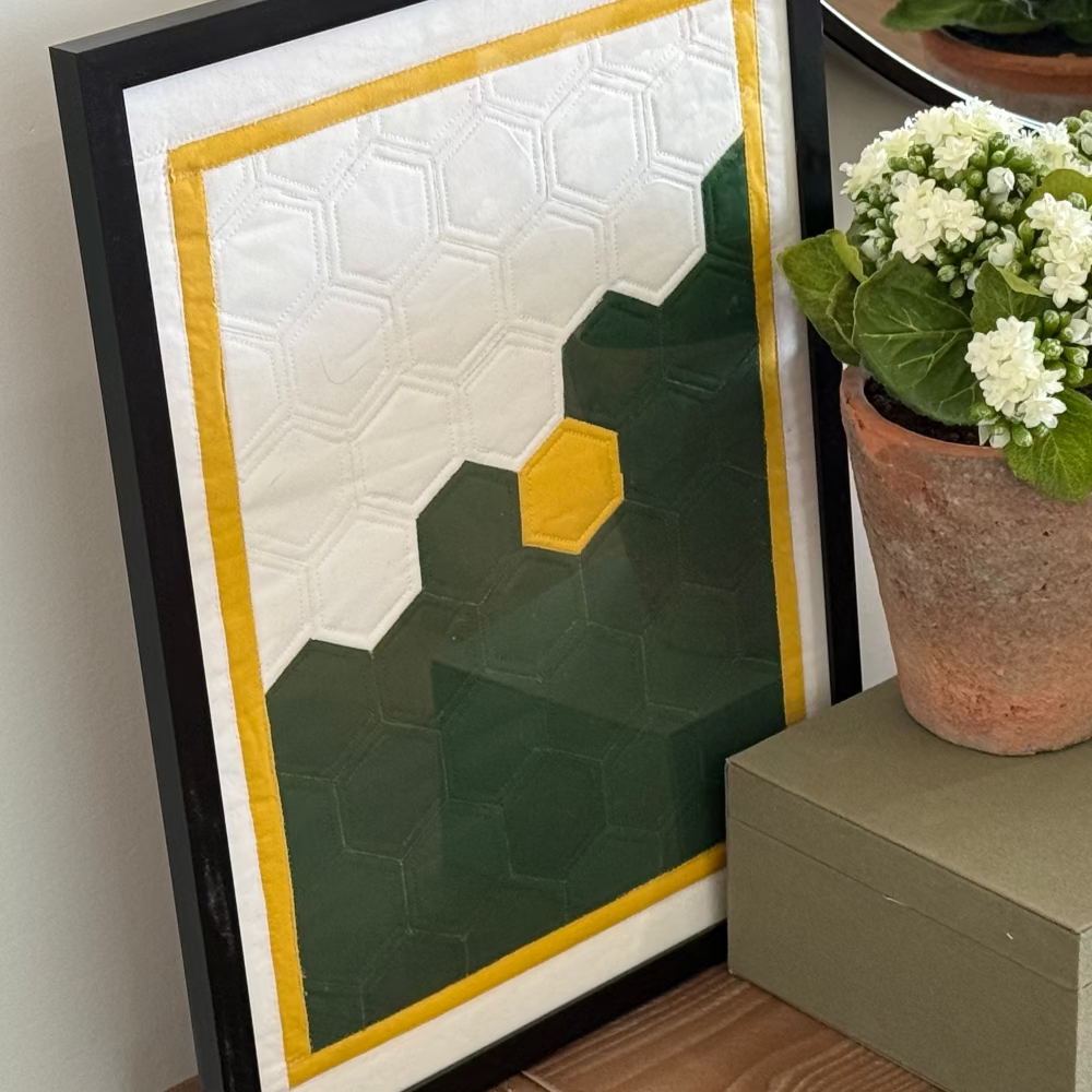 Framed geometric art piece on a shelf with a potted plant and striped container.
