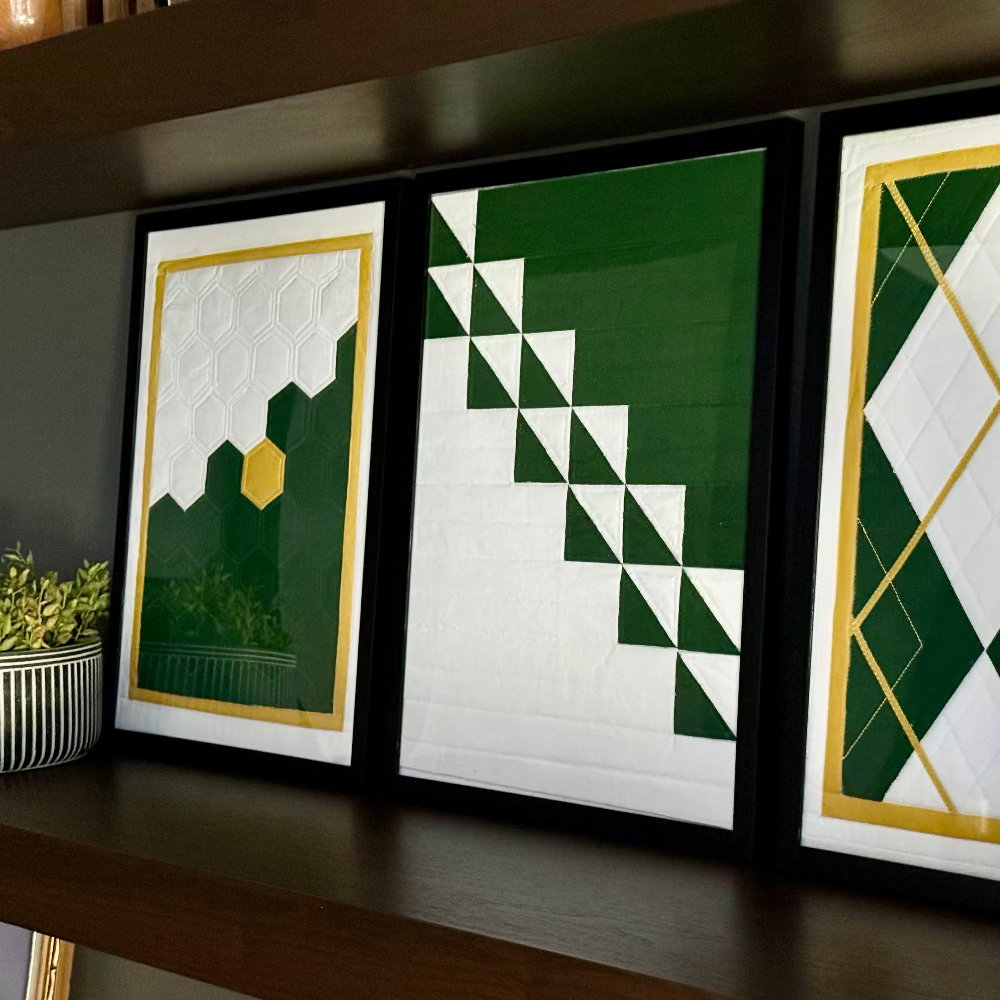 Three framed geometric art pieces on a shelf with a plant and books.