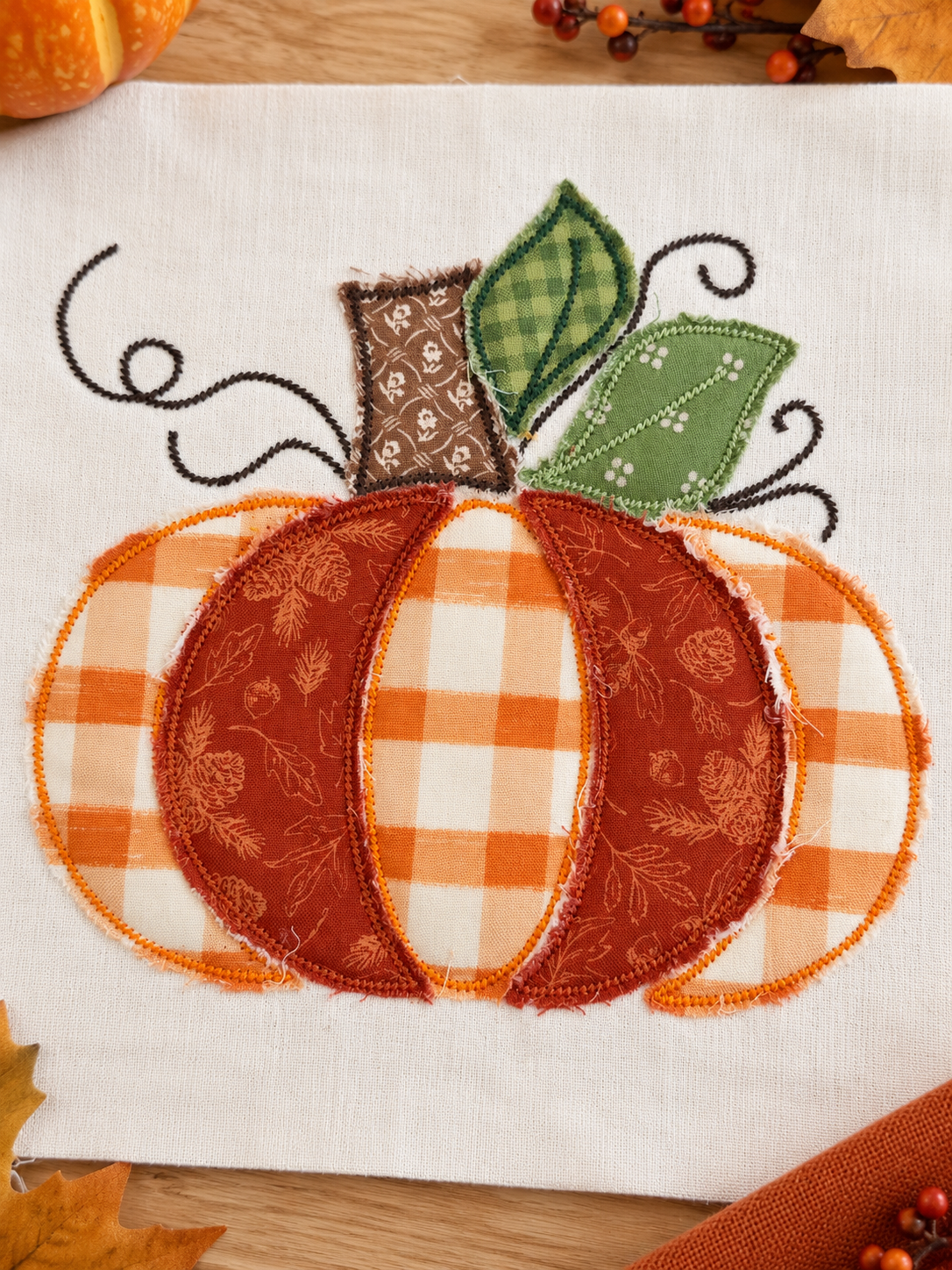 A close-up of a fabric applique pumpkin design on a white textile background, with orange and white gingham sections, a rust-orange patterned center, green leaves, and black vine details. Autumn leaves and small pumpkins are partially visible around the edges.