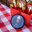 Gold utensils in a red holder on a red and white checkered tablecloth with a patriotic theme.
