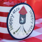 Decorative patch with 'Happy 4th of July' text and rocket design on a red and white checkered tablecloth.