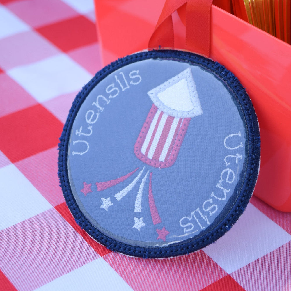 Round utensils holder with American flag design on a red and white checkered tablecloth.