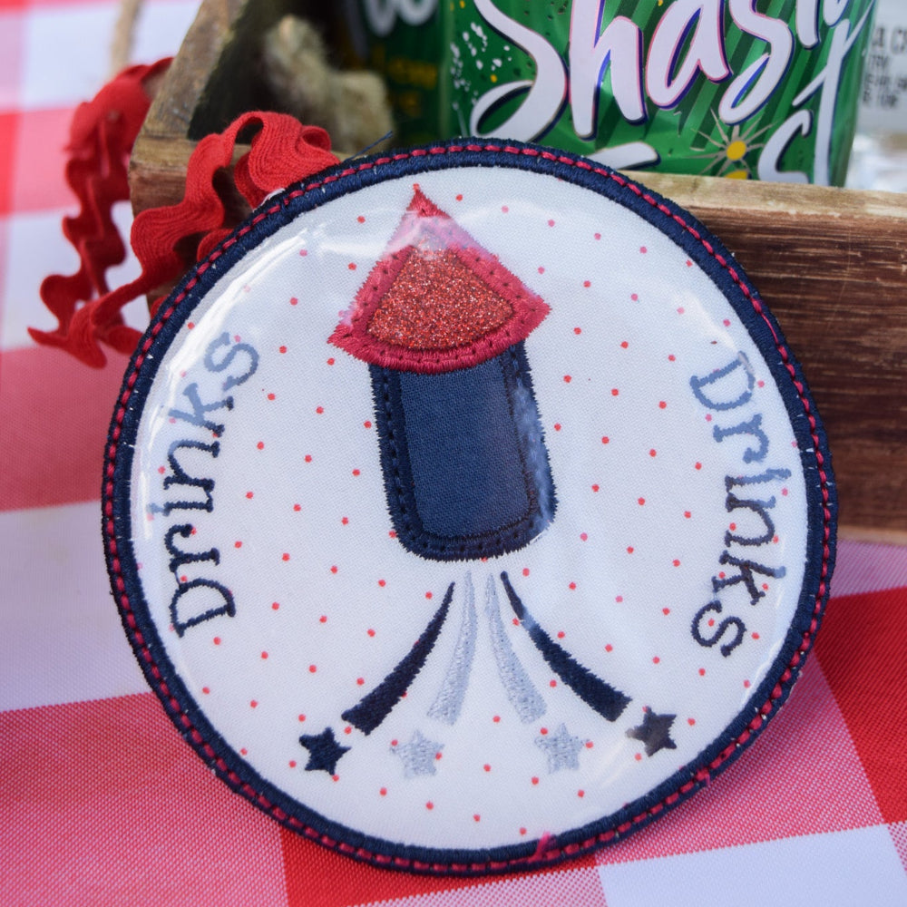 Round patch with a firework design and 'drinks' text on a checkered tablecloth with a can of Shasta in the background.