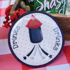 Round patch with a firework design and 'drinks' text on a checkered tablecloth with a can of Shasta in the background.