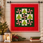 A festive Christmas quilted wall hanging displayed on a black rod against a beige wood-paneled wall. The quilt features a red outer border, a dark green inner field, and green tree and striped pinwheel motifs.