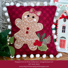 A festive Christmas throw pillow from the Peppermint & Pine Collection, featuring an embroidered gingerbread girl design on a deep red plaid background. The pillow is decorated with white pom-poms around the border and styled with holiday greenery and a small gingerbread house decoration.