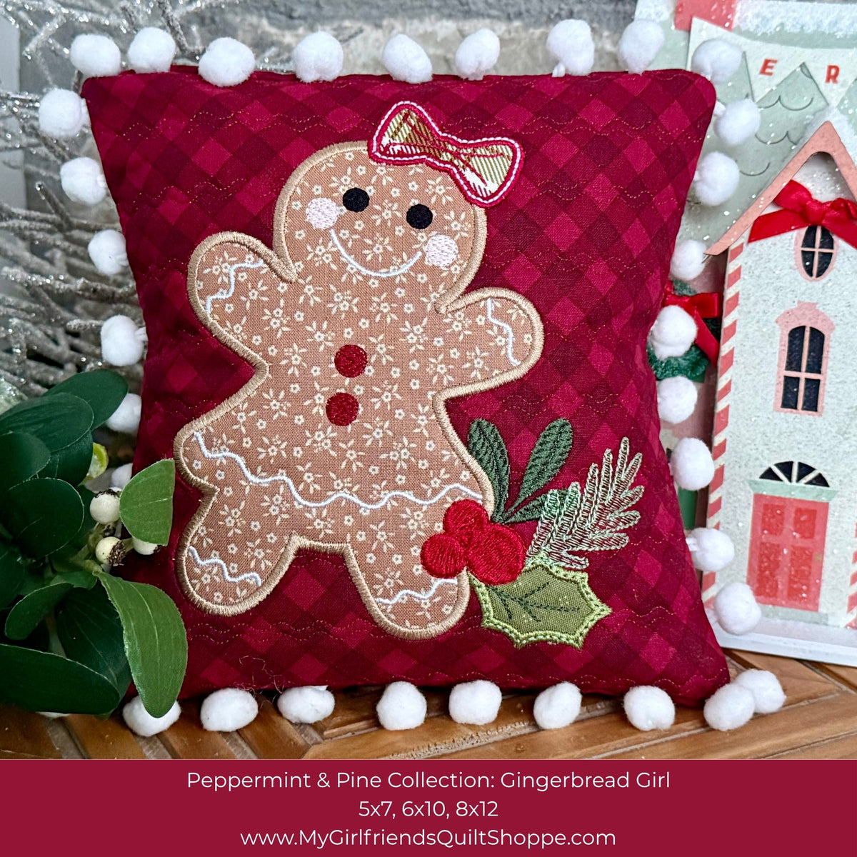A festive Christmas throw pillow from the Peppermint & Pine Collection, featuring an embroidered gingerbread girl design on a deep red plaid background. The pillow is decorated with white pom-poms around the border and styled with holiday greenery and a small gingerbread house decoration.