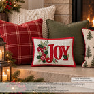 A festive Christmas-themed machine embroidery design preview featuring the word 'JOY' in red with holly and pine branches on a white background, surrounded by decorative throw pillows in a cozy fireplace setting. Text at the bottom specifies dimensions and notes the background quilting is not included.