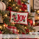A festive embroidered pillow ornament hanging on a decorated Christmas tree. The square off-white fabric ornament has a red gingham border and the word “JOY” in red script, with green holly leaves and red-and-white plaid ribbon accents.