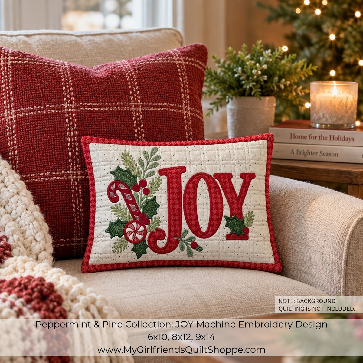 A festive embroidered throw pillow reading “JOY” in red with green holly and candy cane motifs, styled on a beige armchair beside a red plaid pillow in a cozy holiday room setting.