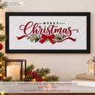 A framed Christmas quilt design featuring the words 'Merry Christmas' in red script over a white dotted background with green pine branches, red berries, and a red bow. The framed print is displayed in a cozy room setting with a lit candle, evergreen branches, and a small house lantern.