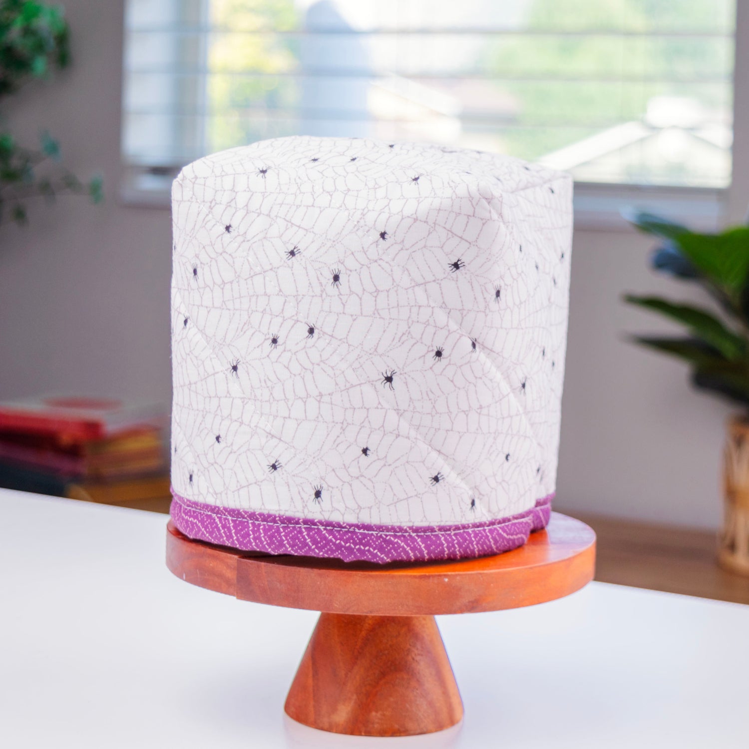 A cylindrical fabric-covered tissue box with a white crackle pattern and tiny spiders, wrapped in a purple binding at the base, displayed on a wooden pedestal stand in a bright indoor setting.