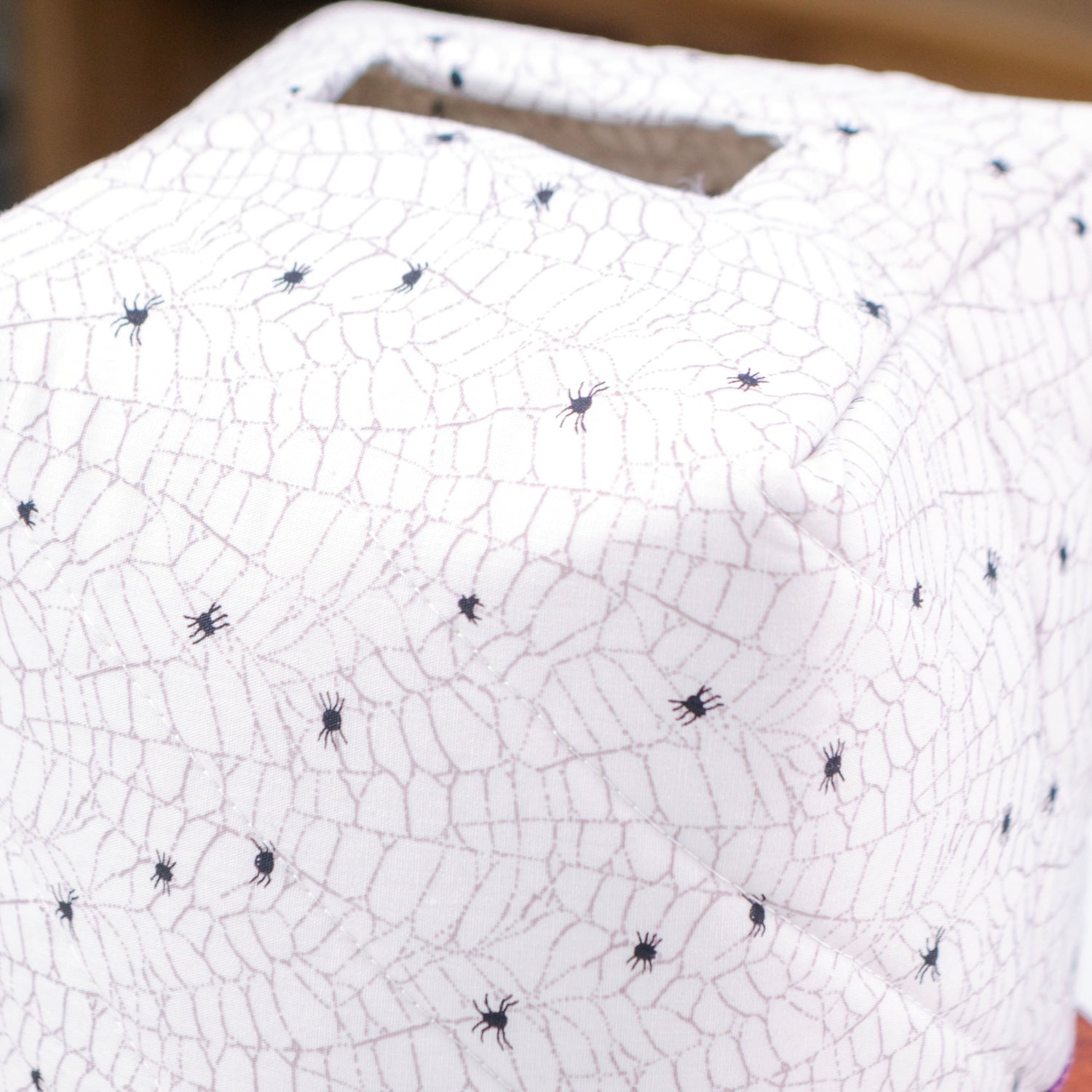A close-up of a white quilted fabric tissue box with a spiderweb and spider print, featuring a purple fabric lining visible along the bottom edge.