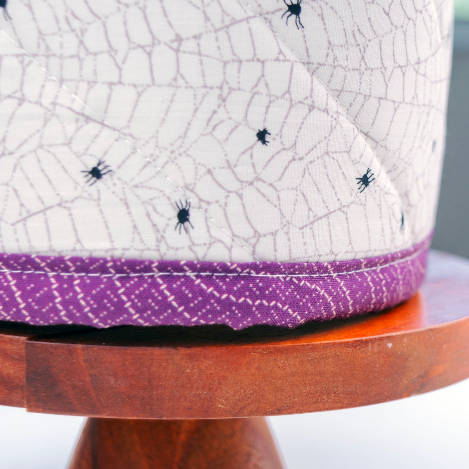 A close-up of a white fabric-covered cylindrical product, likely a speaker or decorative container, featuring a purple patterned band at the base and small black spider motifs. The item is displayed on a round wooden stand against a blurred gray background.