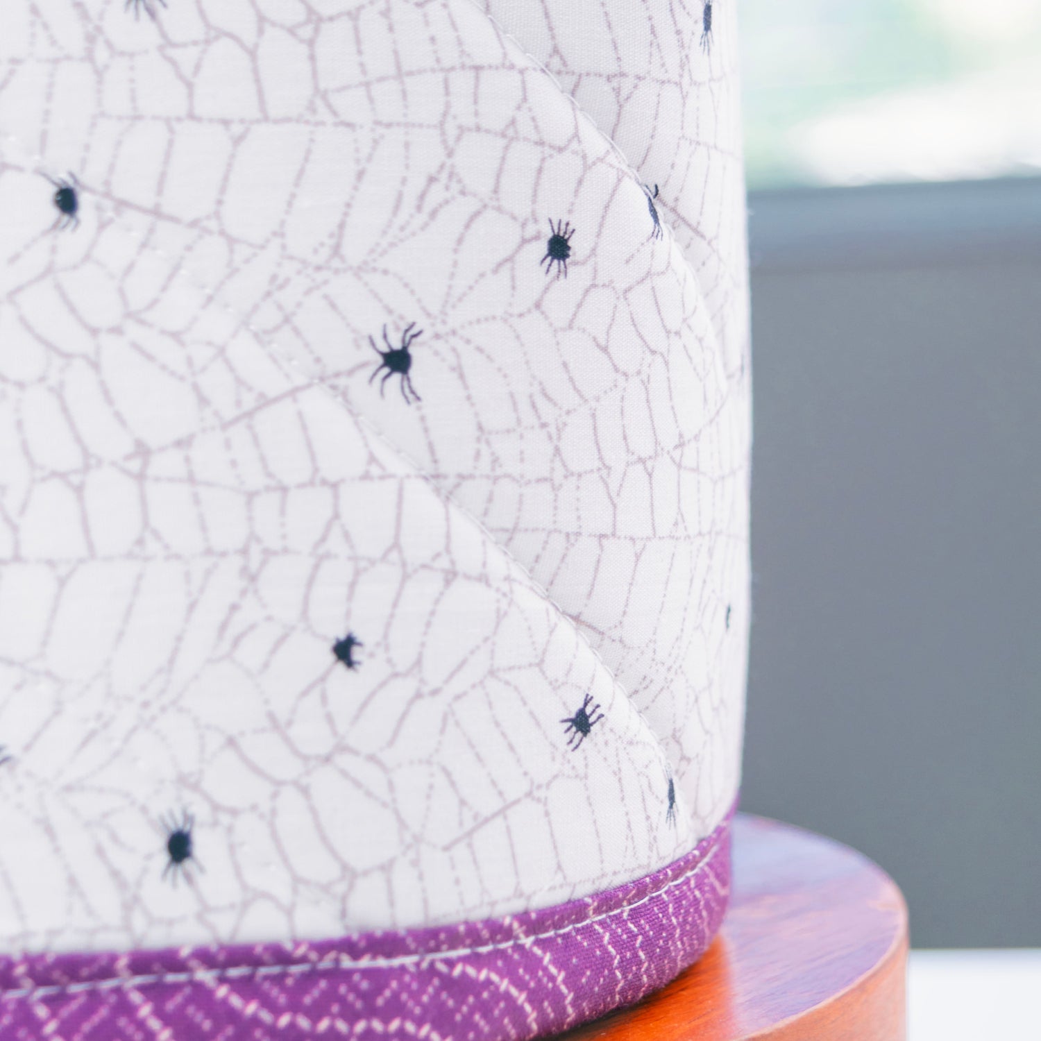 A close-up of a white quilted fabric tissue box, featuring a spiderweb pattern with small black spiders and a purple patterned stitched edge, resting on a wooden stand.