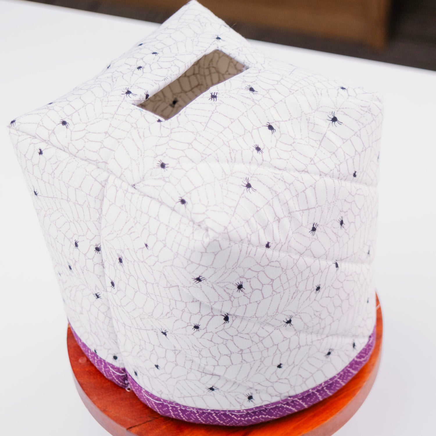 A quilted white tissue box cover with a black spiderweb pattern and small black spider motifs, displayed on a wooden stand on a white table.