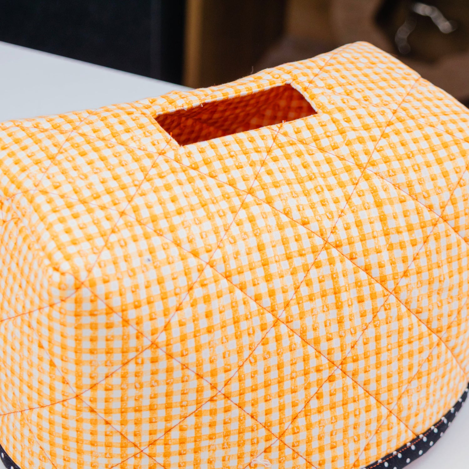 A quilted fabric tissue box cover with a yellow and white gingham pattern, featuring a rectangular opening at the top and a black polka-dot trim along the base, shown on a white tabletop.
