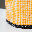 A close-up of a quilted fabric ottoman or pouf with a bright orange and white gingham pattern, finished with a black band of white polka dots along the base.