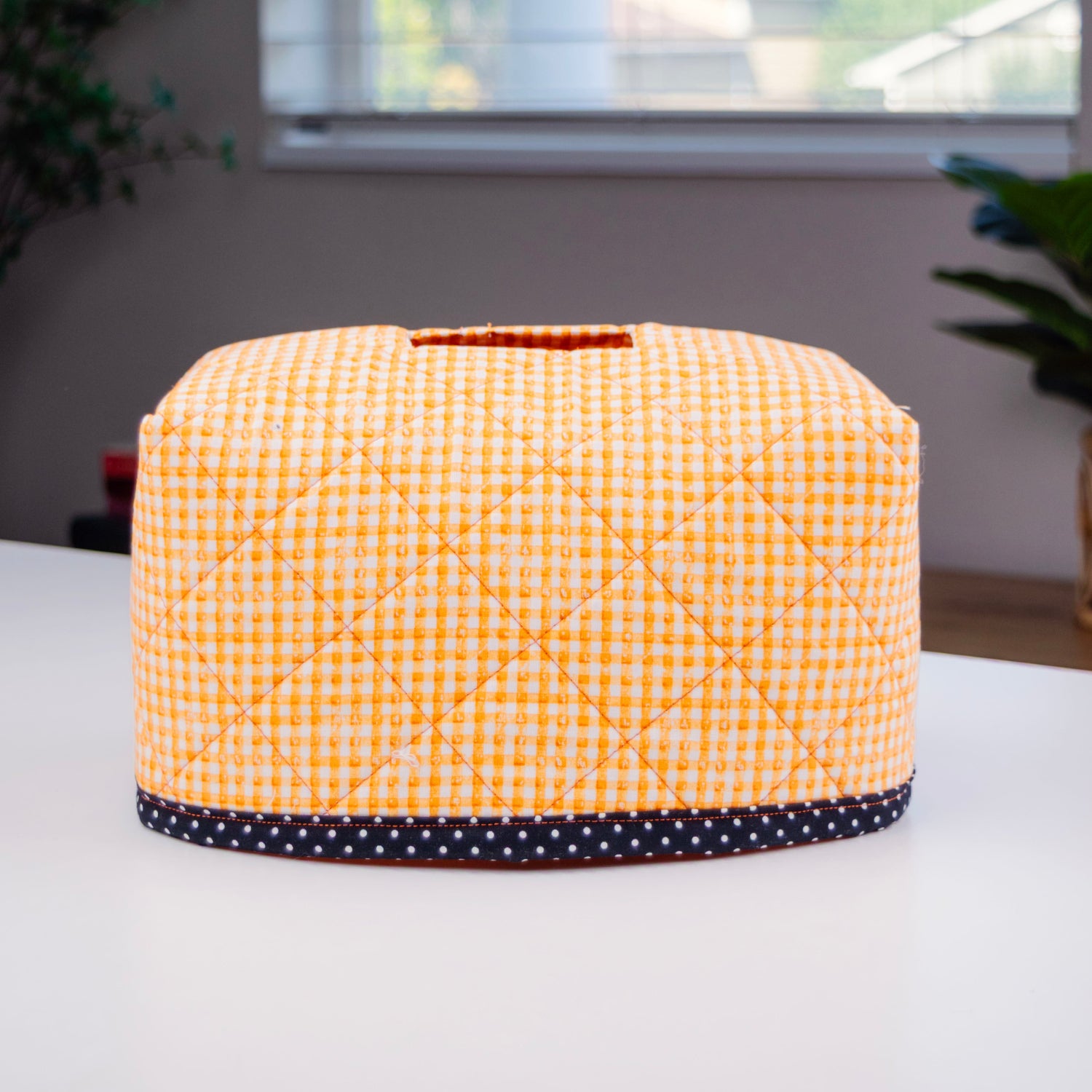 A quilted fabric tissue box cover with a yellow and white gingham pattern and a navy blue polka-dot trim, sitting on a white table in front of a window.