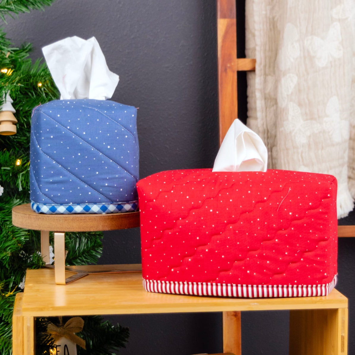 Two quilted fabric tissue box covers, one blue with white dots and one red with white dots, displayed on a wooden shelf next to a Christmas tree and curtain.