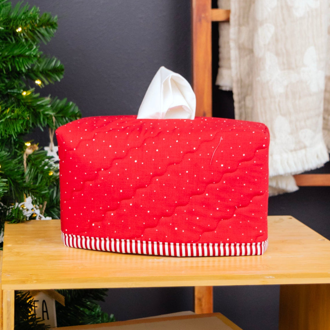 A red quilted tissue box cover with a white tissue peeking out, placed on a light wood shelf next to a decorated Christmas tree.