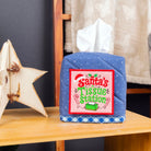 A quilted blue tissue box cover with a white tissue protruding, decorated with a red and green 'Santa's Tissue Station' patch. It sits on a wooden table next to a wooden star ornament, with a Christmas tree and a chair in the background.
