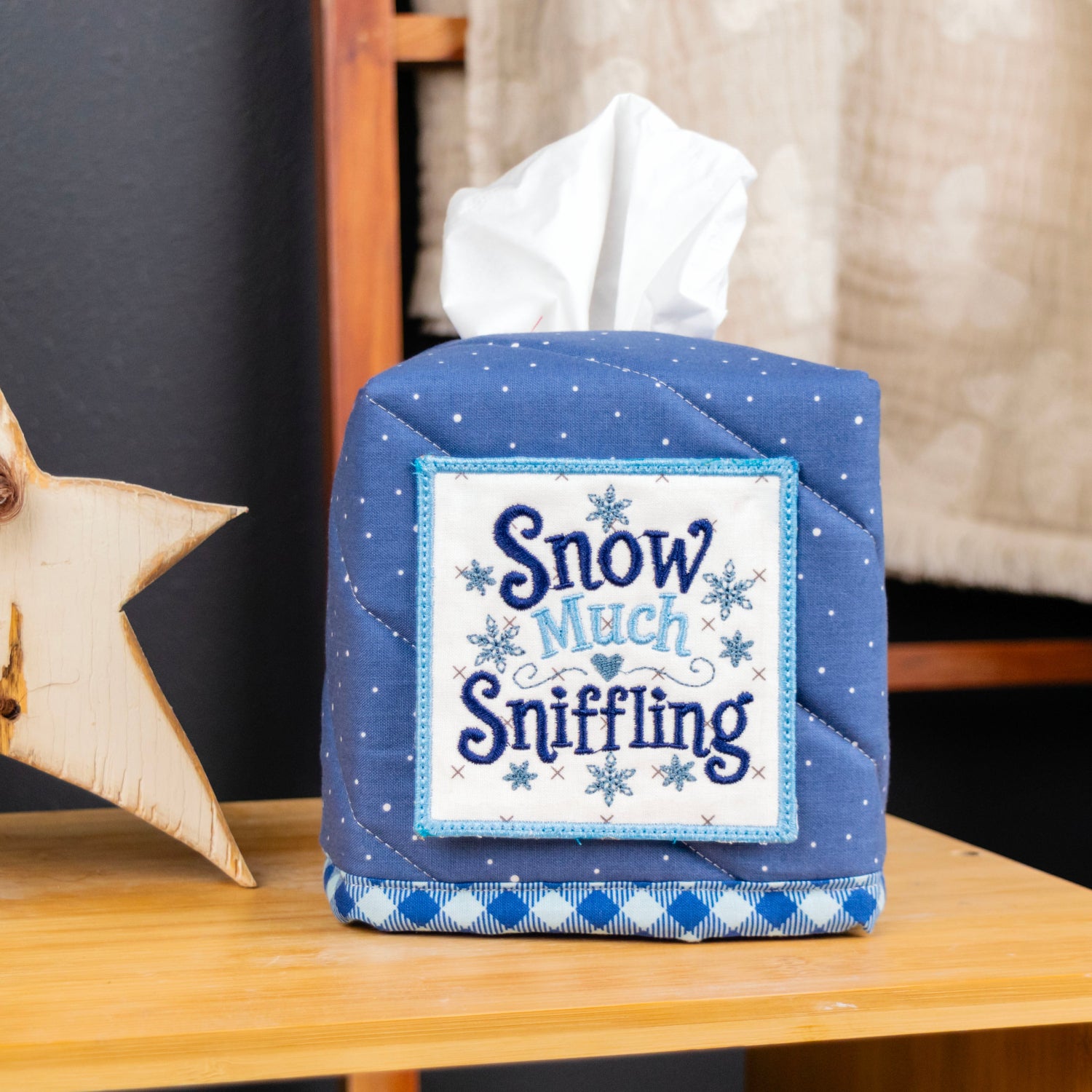 A quilted blue fabric tissue box cover with a white tissue insert, featuring a front patch that reads 'Snow Much Sniffing' with snowflake details. The cover is styled on a wooden table next to a wooden star ornament.