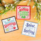 Three embroidered Christmas-themed square patches displayed on a wooden surface with decorative evergreen branches. The patches read 'Santa's Tissue Station,' 'Seasons Sneezings,' and 'Snow Much Sniffling.